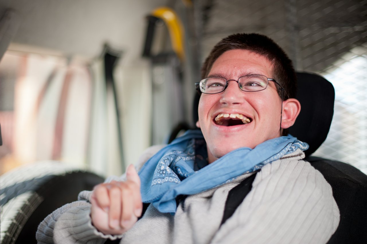 A young white man with cerebral palsy sites in a wheelchair in his workshop. He has brown hair and wears glasses, a white sweater, and a blue handkerchief around his neck.