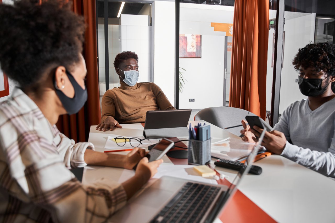 A Black woman, a Black man, and a Latino man sit around a conference table in a contemporary office space. All three wear protective masks and work on their laptops.