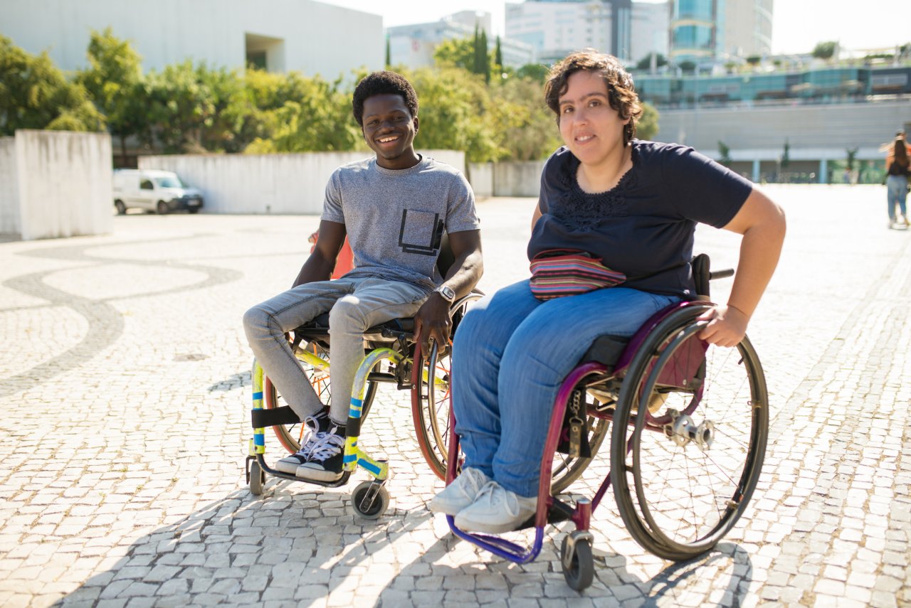 An interracial couple with disabilities sit next to each other outside in the sunshine, smiling at the camera.