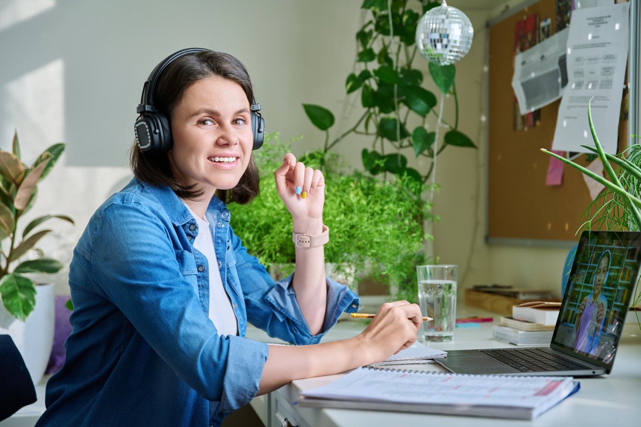 A white woman with short brown hair sits at her desk, attending a virtual meeting. She wears a blue denim shirt and has on over-ear headphones to assist with a hearing disability.