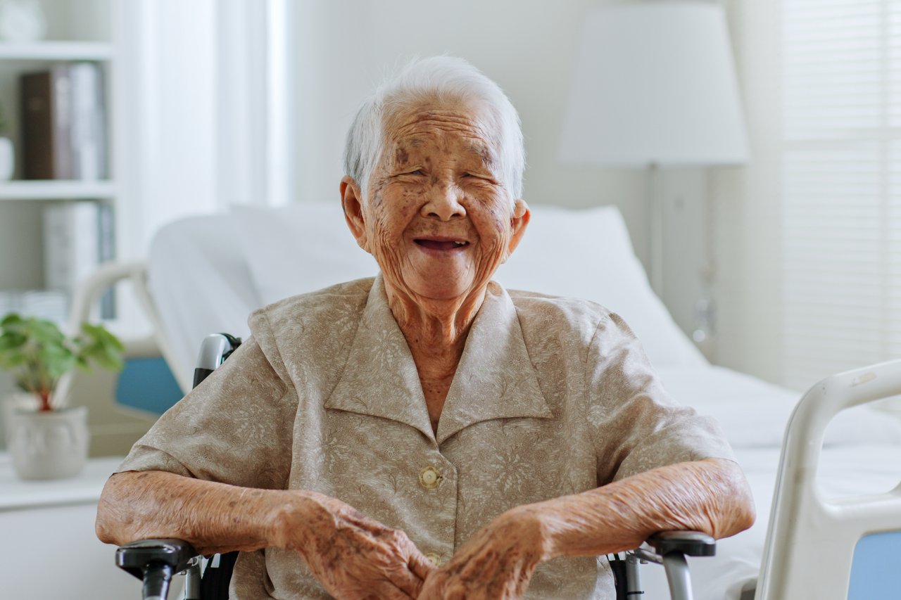 A senior Asian woman sits inside her house, in front of a hospital bed. She has short white hair and sits in a wheelchair.