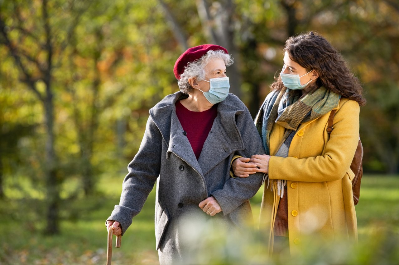 A senior woman wearing a mask and a heavy coat walks outside with her daughter, a young white woman with brown hair who also wears a mask.