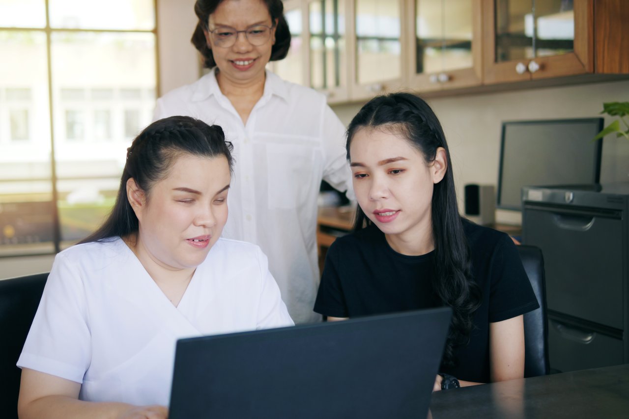 Two Asian women stand together by the computer, which is used by a blind Asian woman.